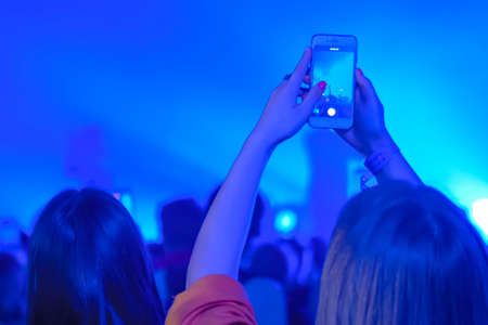 Teenager taking photo or recording video of live music concert with smartphone - back view. Bright colorful blue stage lighting. Nightlife, technology conceptの写真素材
