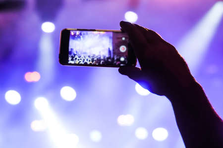 Man hands silhouette taking photo or recording video of live music concert with smartphone in front of stage of nightclub - close up. Photography, entertainment, technology conceptの写真素材