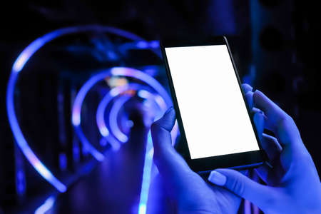 Mockup image: woman hands using smartphone device with white display at interactive exhibition or museum with blue illumination - close up. Mock up, entertainment, white screen, technology conceptの写真素材