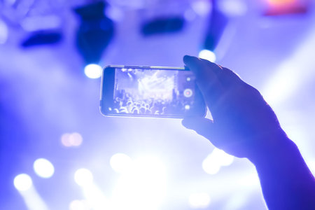 Man hands silhouette taking photo or recording video of live music concert with smartphone in front...