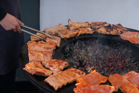 Chef holding tongs and grilling fresh juicy meat steaks on brazier at summer local food market -...