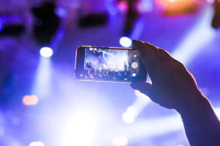 Man hands silhouette taking photo or recording video of live music concert with smartphone in front of stage of nightclub - close up, lens flare. Photography, entertainment, technology conceptの写真素材
