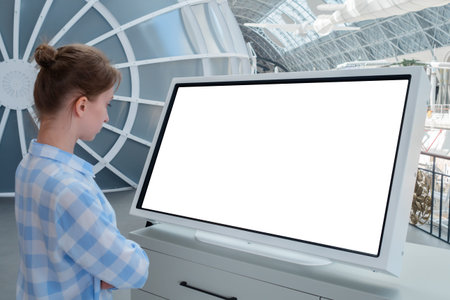 Woman in blue plaid shirt looking at blank interactive touchscreen white display of electronic kiosk at futuristic exhibition or museum. White screen, mock up, copyspace, template, technology concept.の写真素材