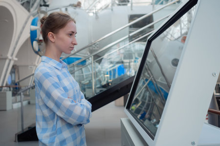 Portrait of woman in blue plaid shirt looking at interactive touchscreen display of electronic kiosk...