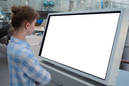 Woman in blue plaid shirt looking at blank interactive touchscreen white display of electronic kiosk at futuristic exhibition or museum. White screen, mock up, copyspace, template, technology concept.の写真素材