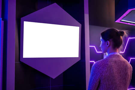 Woman looking at blank digital interactive white display wall at exhibition or museum with purple futuristic scifi interior. White screen, mock up, future, copyspace, template, technology conceptの写真素材