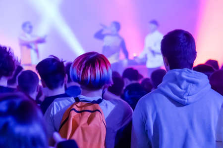 Teenagers partying, cheering, raising hands up and jumping at rock concert in front of stage of nightclub - back view. Bright colorful stage lighting. Nightlife and entertainment conceptの写真素材