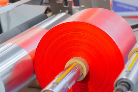 Moving roller with flat polyethylene red film - automatic plastic bag making machine at exhibition: close up side view. Recycling, manufacturing, industry, automated technology equipment conceptの写真素材