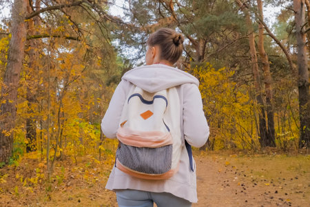 Back view of young woman with backpack walking in autumn park, forest. Active outdoor lifestyle,...