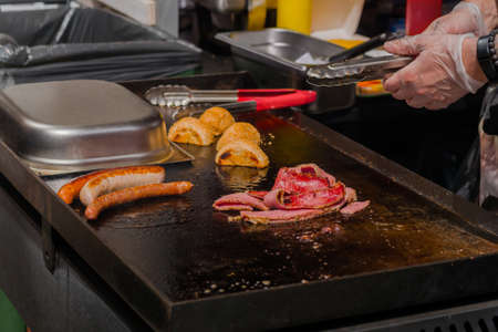 Chef preparing burger buns, bacon slices and long sausages on grill at summer local food market - close up view. Outdoor cooking, gastronomy, cookery, street food conceptの写真素材