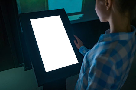 Woman looking at white blank interactive touchscreen display of electronic multimedia kiosk in dark...