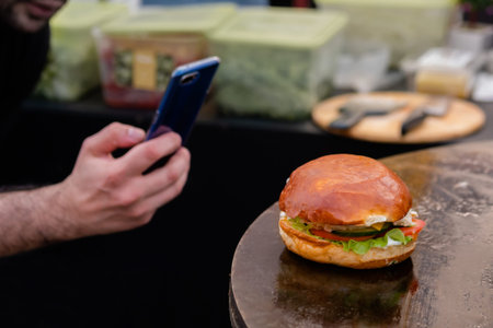 Man hand holding smartphone, taking photo of burger with fish on brazier at summer local food market...