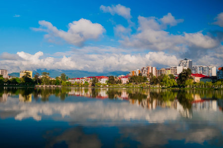 View of the blue sky with white clouds over modern apartment, residential buildings with reflection in the water surface. 6 May Park on the shore of lake in Batumi in the afternoonの写真素材