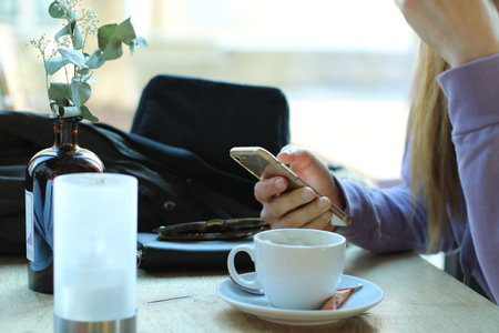 The woman is sitting in the cafe. The woman is drinking coffee and the woman is holding a mobile phone.の写真素材