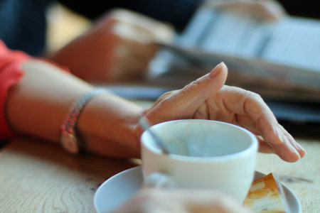 The old woman is sitting in the cafe. There is a white coffee cup on the table. Woman is holding coffee cup in her hand. The picture shows a woman's hand and a coffee cup.の写真素材