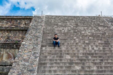 A Chinese tourist sitting on steps in the ruins of the architecturally significant Mesoamerican pyramids and green grassland located at at Teotihuacan, an ancient Mesoamerican city located in a sub-valley of the Valley of Mexicoのeditorial素材