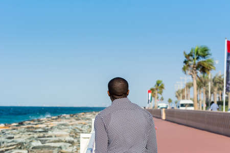 An African man watching the seascape at the crescent road and the breakwater and stones and background of Persian gulf in the Palm Jumeirah island in Dubai of the United Arab Emirates.の写真素材