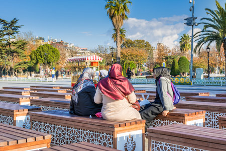 Turkish women wearing traditional clothes and sitting on the chairs located between Blue Mosque and Hagia Sophia mosque under sunlight in the morning in autumn in Istanbul, Turkeyのeditorial素材