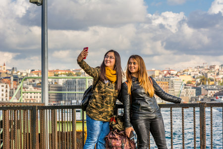 Two blond beautiful Turkish girls taking selfies with cellphone at the Bosphorus strait on a sunny day with background cloudy sky, Istanbul, Turkey.のeditorial素材