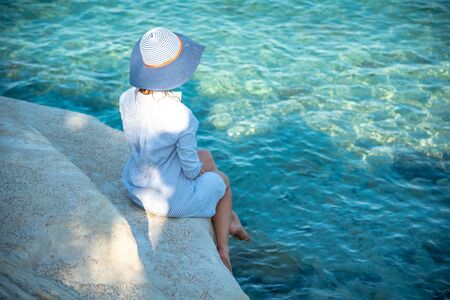 Carefree happy woman is enjoying blue sea, sitting on stone with view from above. A girl in the hat is sitting on a rock with her back. Freedom concept. Travel and relax.の写真素材