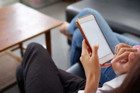 Young woman using black tablet device with white screen. Woman holding tablet, scrolling pages while sitting on the sofa in the living room.の写真素材