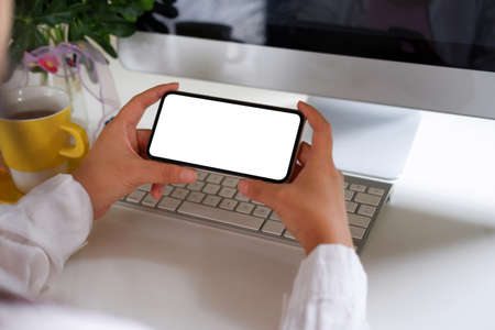 Woman using mobile phone, blank screen on a white desk with a coffee cup, keyboard, mouse, flowers.の写真素材