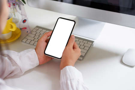 Woman using mobile phone, blank screen on a white desk with a coffee cup, keyboard, mouse, flowers.の写真素材