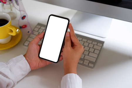 Woman using mobile phone, blank screen on a white desk with a coffee cup, keyboard, mouse, flowers.の写真素材