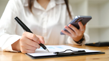 Asian woman writing and using smart phone at workplace.の写真素材