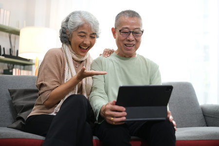 senior couple sitting on sofa using tablet while video call online with family in living room at...