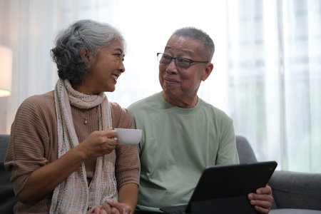 senior couple sitting on sofa using tablet while video call online with family in living room at home.の写真素材