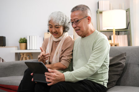 senior couple sitting on sofa using tablet while video call online with family in living room at home.の写真素材