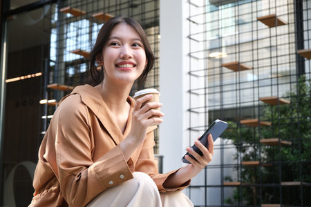 Portrait of cheerful young female holding coffee cup and using mobile mobile while sitting in coffee shop.の写真素材