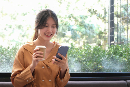 Portrait of cheerful young female holding coffee cup and using mobile mobile while sitting in coffee shop.の写真素材