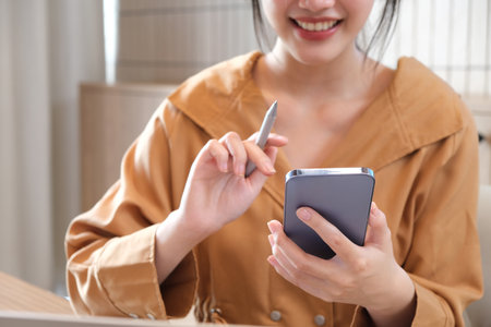 Smiling and charming Asian woman sit at her desk with a smartphone playing social media applications and transacting at her desk.の写真素材