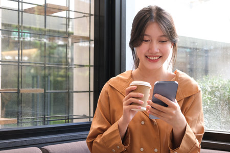 Portrait of cheerful young female holding coffee cup and using mobile mobile while sitting in coffee shop.の写真素材