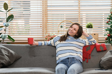 Beautiful caucasian woman drinking coffee and using smartphone on sofa near decorated Christmas tree..の写真素材