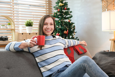 Beautiful caucasian woman drinking coffee and using smartphone on sofa near decorated Christmas tree..の写真素材