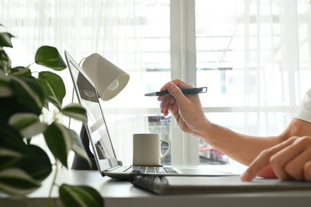 A person is writing on a laptop with a pen. The scene is set in a room with a potted plant and a cup.の写真素材