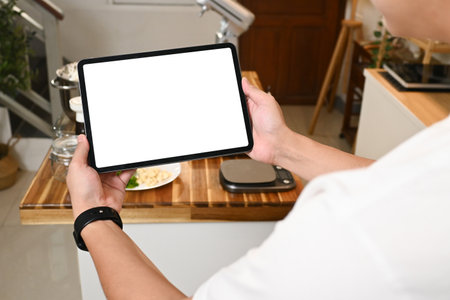 A person is holding a tablet in a kitchen with a lot of utensils and bowls. The tablet is empty, and the kitchen appears to be a place where someone is preparing food..の写真素材