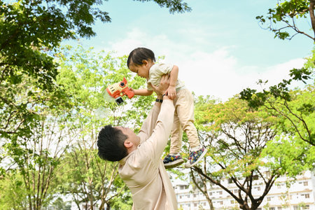 A man is holding a small child in the air. The scene is set in a park with trees in the background. Scene is playful and joyful.の写真素材