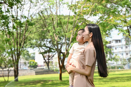 A woman is holding a baby in a park. The woman is looking at the baby with a smile on her face. The park is filled with trees and grass, creating a peaceful.の写真素材