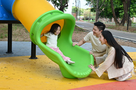 A family is standing in front of a slide. The child is sitting on the slide while the parents look on. Scene is happy and playful..の写真素材