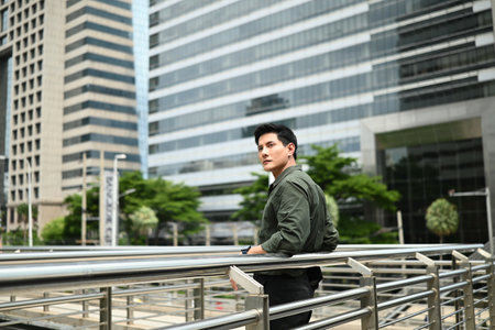 A man is smiling and leaning on a railing in front of a large building. The man is enjoying the view of the city.の写真素材