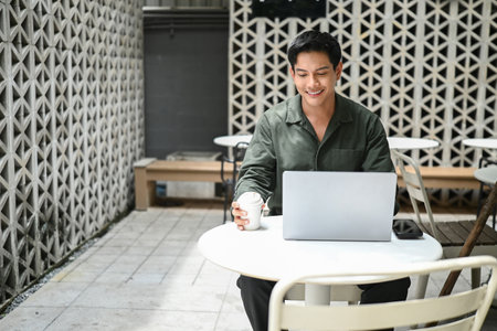 A man is sitting outside at a small table, deeply engrossed in his laptop while also enjoying a cup of coffee.の写真素材