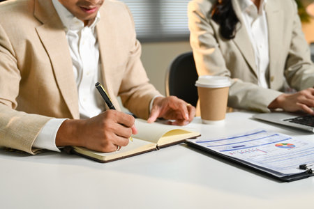 Two people in suits are sitting at a desk with a notebook and a laptop. One of them is writing in the notebook while the other is typing on the laptop..の写真素材