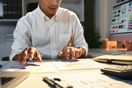 A man is sitting at a desk with a computer monitor and a tablet. He is looking at a piece of paper and seems to be working on a project.の写真素材