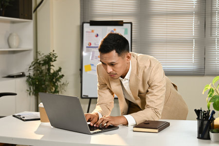 A man in a suit is sitting at a desk with a laptop open in front of him. He is typing on the keyboard and he is focused on his work.の写真素材