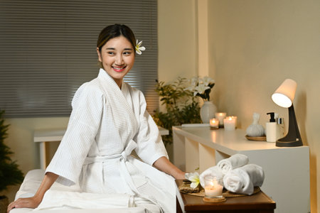 A woman is sitting on a bed in a spa, wearing a white robe and smiling. The room is decorated with flowers and candles, creating a relaxing atmosphere.の写真素材