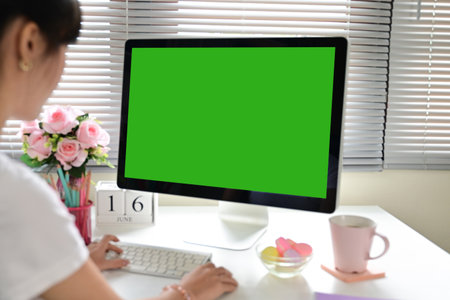 A woman is sitting at a desk with a green computer monitor in front of her. The monitor is blank, and the woman is typing on a keyboard. The room has a floral arrangement on the desk and a pink cupの写真素材